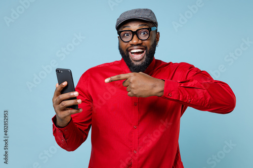 Excited young bearded african american man 20s wearing casual red shirt eyeglasses cap pointing index finger on mobile cell phone typing sms message isolated on pastel blue background studio portrait. © ViDi Studio