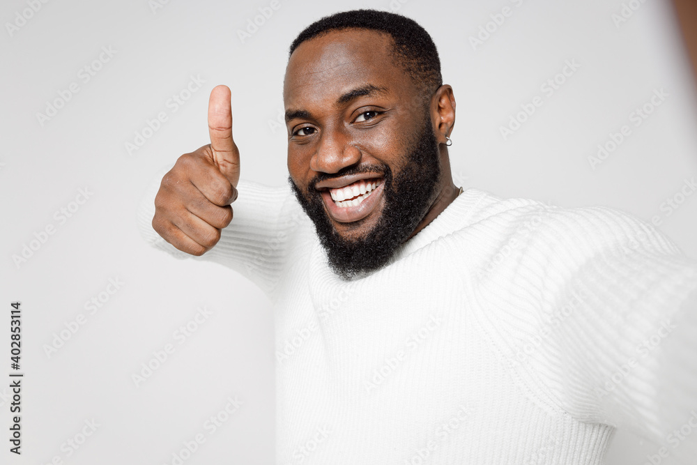 Fototapeta premium Close up of smiling young african american man 20s in casual basic sweater doing selfie shot on mobile phone showing thumb up looking camera isolated on white color wall background studio portrait.