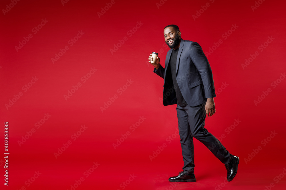 Full length side view of smiling young african american business man 20s in classic jacket suit hold paper cup of coffee or tea looking camera isolated on bright red color background studio portrait. Full length side view of smiling young african american business man 20s in classic jacket suit hold paper cup of coffee or tea looking camera isolated on bright red color background studio portrait.
