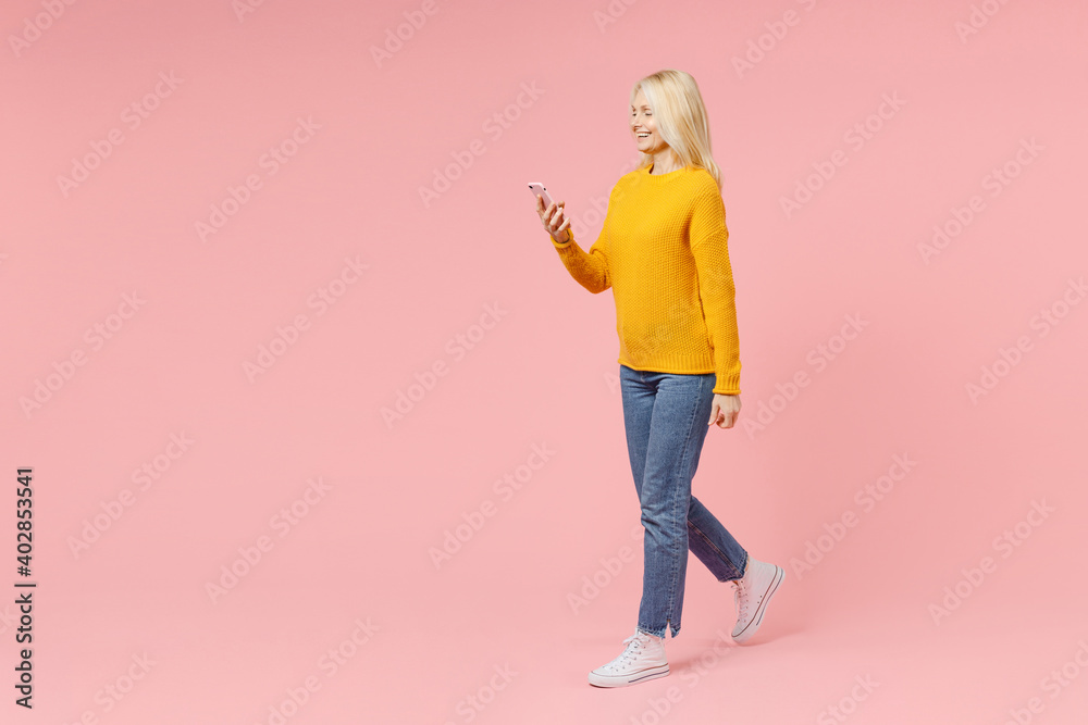 Full length side view of smiling elderly gray-haired blonde woman lady 40s 50s years old in yellow sweater using mobile cell phone typing sms message isolated on pink color background studio portrait. Full length side view of smiling elderly gray-haired blonde woman lady 40s 50s years old in yellow sweater using mobile cell phone typing sms message isolated on pink color background studio portrait.
