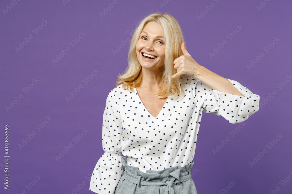 Excited funny elderly gray-haired blonde woman lady 40s 50s years old wearing white dotted blouse doing phone gesture like says call me back isolated on bright violet color background studio portrait. Excited funny elderly gray-haired blonde woman lady 40s 50s years old wearing white dotted blouse doing phone gesture like says call me back isolated on bright violet color background studio portrait.