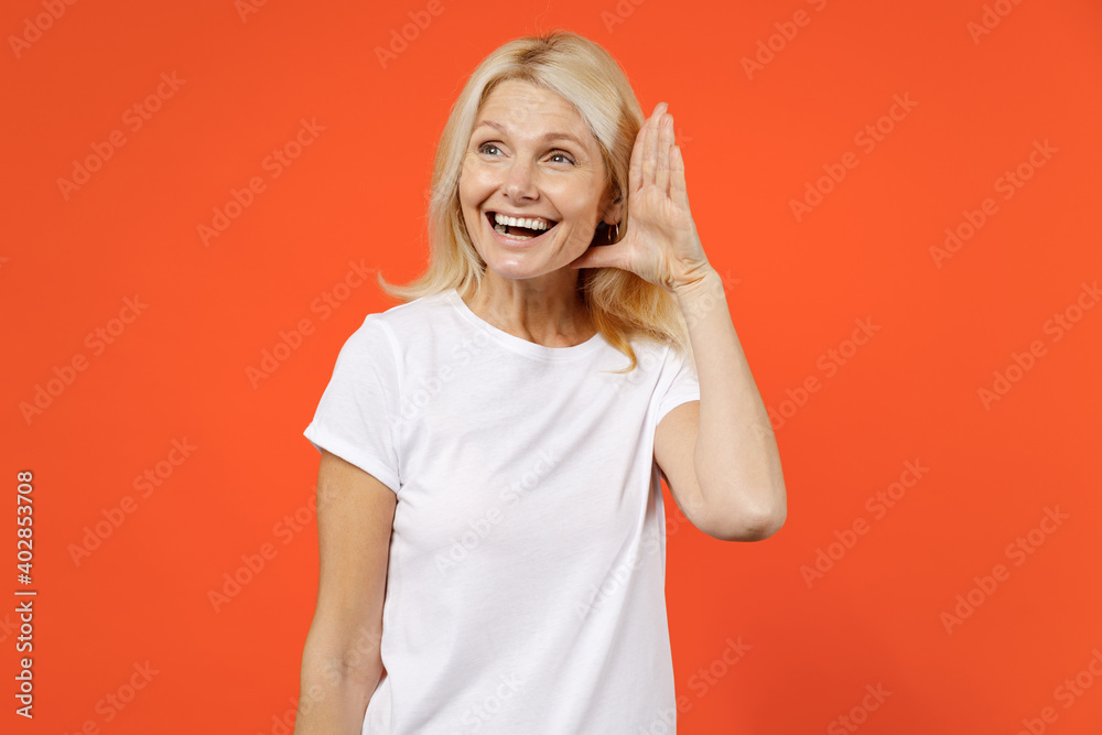 Funny curious elderly gray-haired blonde woman lady 40s 50s years old in white casual t-shirt standing try to hear you overhear listening intently isolated on orange color background studio portrait. Funny curious elderly gray-haired blonde woman lady 40s 50s years old in white casual t-shirt standing try to hear you overhear listening intently isolated on orange color background studio portrait.