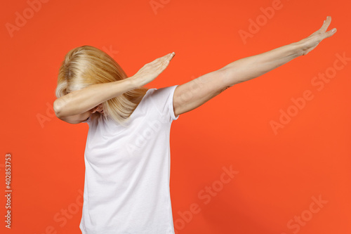 Cheerful elderly gray-haired blonde woman lady 40s 50s in white basic t-shirt doing dab hip hop dance hands gesture youth sign hiding covering face isolated on orange color background studio portrait.