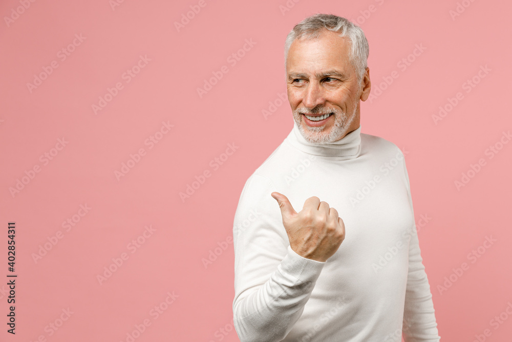 Side view of smiling elderly gray-haired mustache bearded man wearing casual basic white turtleneck pointing thumb aside on mock up copy space isolated on pastel pink color background studio portrait. Side view of smiling elderly gray-haired mustache bearded man wearing casual basic white turtleneck pointing thumb aside on mock up copy space isolated on pastel pink color background studio portrait.