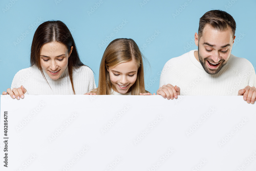 Excited young parents mom dad with child kid daughter teen girl in sweaters hold big white empty blank billboard mock up copy space isolated on blue background studio. Family day parenthood concept. Excited young parents mom dad with child kid daughter teen girl in sweaters hold big white empty blank billboard mock up copy space isolated on blue background studio. Family day parenthood concept.