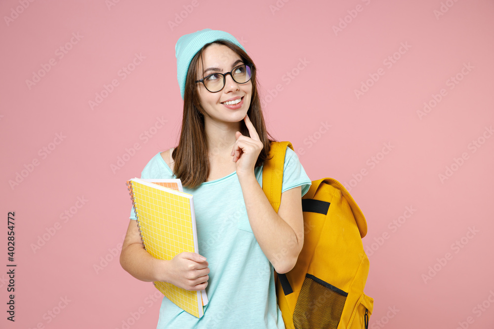 Pensive woman student in blue t-shirt hat eyeglasses backpack hold notebooks put hand prop up on chin isolated on pink background studio portrait. Education in high school university college concept.