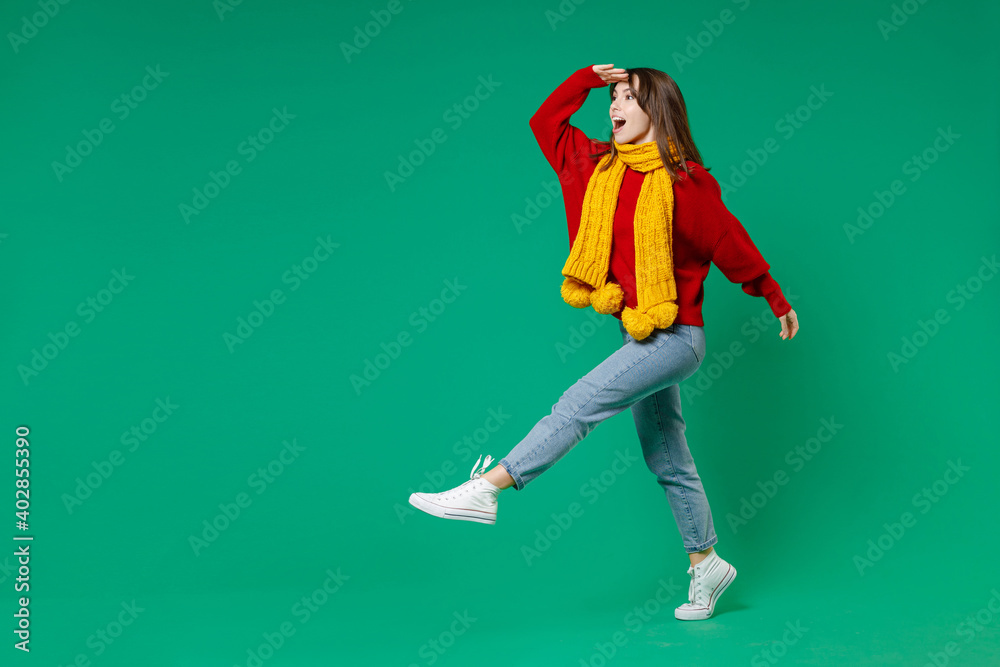 Full length side view of excited shocked young brunette woman 20s in knitted red sweater yellow scarf holding hand at forehead looking far away distance isolated on green background studio portrait.
