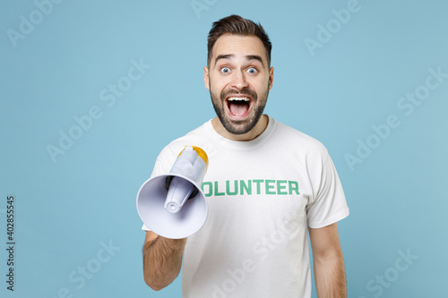 Surprised excited shocked young man wearing white volunteer t-shirt screaming in megaphone isolated on blue color background studio portrait. Voluntary free work assistance help charity grace concept.