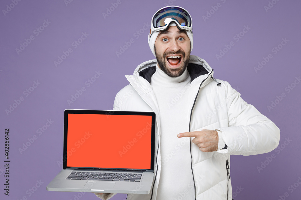 Amazed skier man in white jacket ski goggles mask point index finger laptop computer with blank empty screen spend weekend in mountains isolated on purple background. People lifestyle hobby concept. Amazed skier man in white jacket ski goggles mask point index finger laptop computer with blank empty screen spend weekend in mountains isolated on purple background. People lifestyle hobby concept.