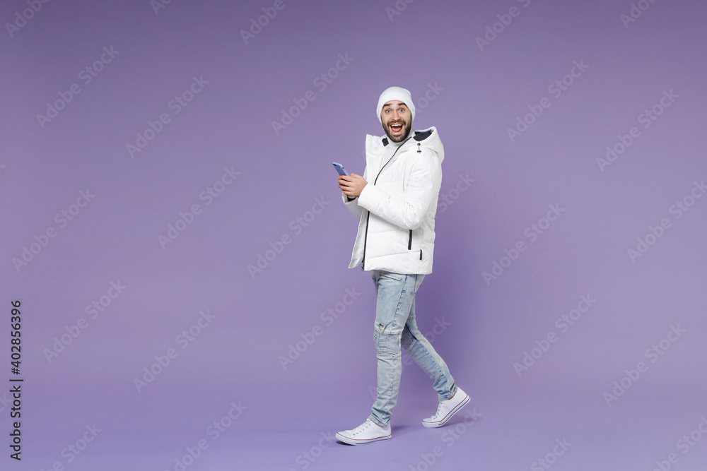 Full length side view surprised man in warm white windbreaker jacket hat using mobile cell phone typing sms message isolated on purple background studio portrait. People lifestyle cold season concept. Full length side view surprised man in warm white windbreaker jacket hat using mobile cell phone typing sms message isolated on purple background studio portrait. People lifestyle cold season concept.