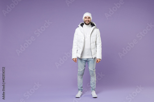 Full length of smiling attractive man in warm white padded windbreaker jacket hat standing looking camera isolated on purple background studio portrait. People lifestyle cold winter season concept.