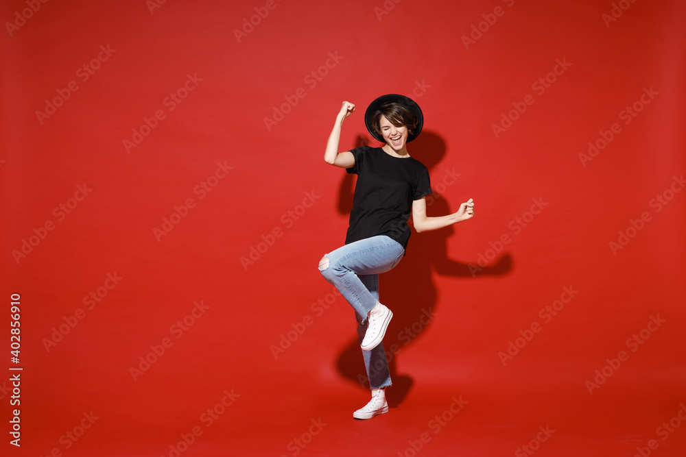 Full length of happy joyful young brunette woman 20s in casual basic black t-shirt hat doing winner gesture celebrating clenching fists say yes isolated on bright red color background studio portrait.