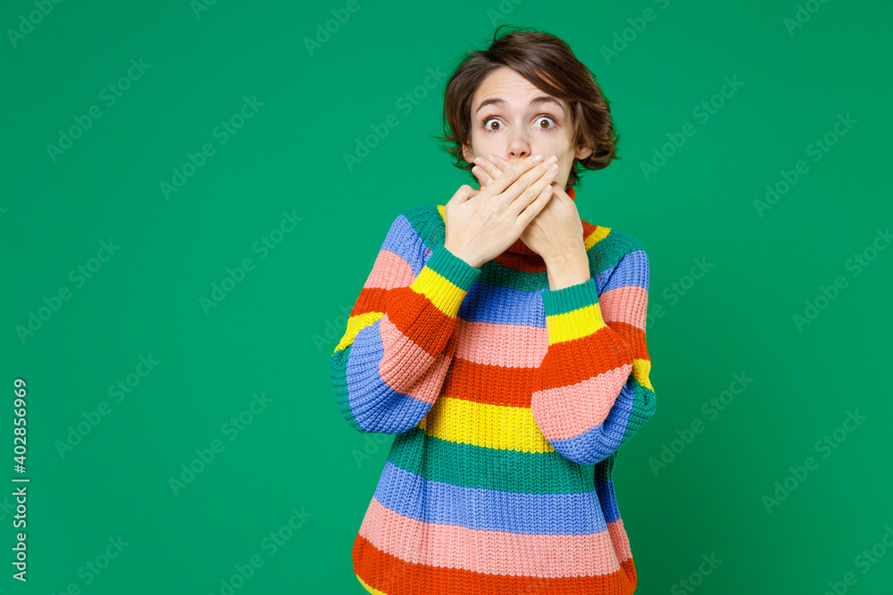 Shocked amazed young brunette woman 20s years old wearing basic casual colorful sweater standing covering mouth with hands looking camera isolated on bright green color background studio portrait. Shocked amazed young brunette woman 20s years old wearing basic casual colorful sweater standing covering mouth with hands looking camera isolated on bright green color background studio portrait.