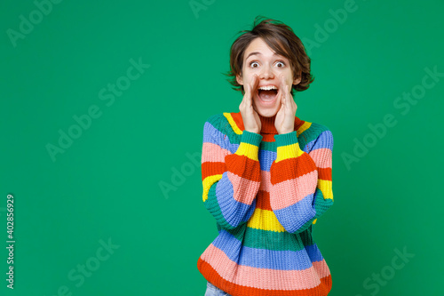 Surprised shocked young brunette woman 20s wearing casual basic colorful sweater standing scream news with hands near mouth looking camera isolated on bright green color background studio portrait.