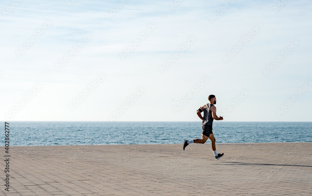 Fit young man running along seafront. Male runner jogging early in
