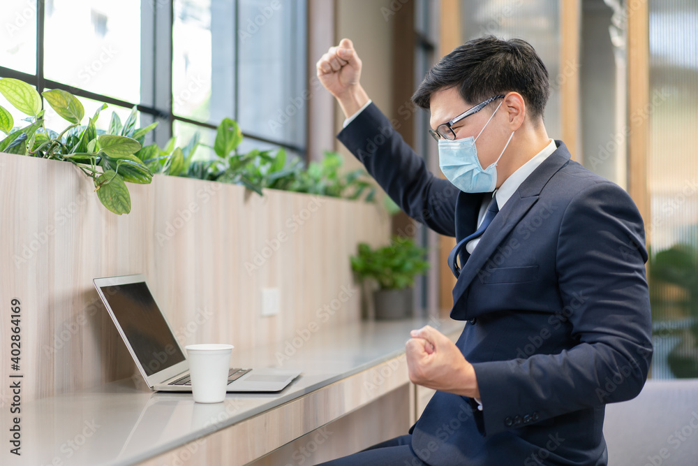 Asian businessman in suit and wearing medical mask protective raising his arm up to celebrate work success achievement while looking at laptop at the workplace Asian businessman in suit and wearing medical mask protective raising his arm up to celebrate work success achievement while looking at laptop at the workplace