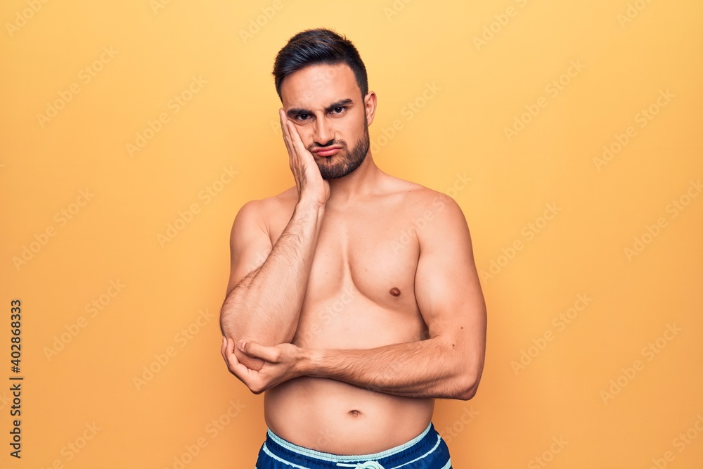 Young handsome man with beard wearing sleeveless t-shirt standing over yellow background thinking looking tired and bored with depression problems with crossed arms.