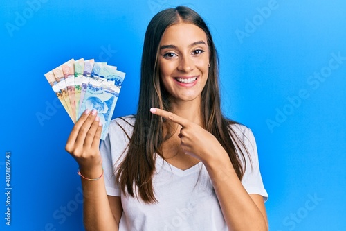 Young hispanic woman holding canadian dollars smiling happy pointing with hand and finger photo