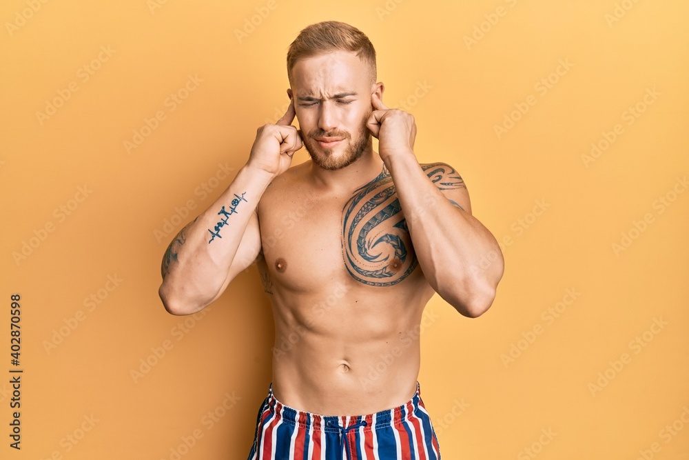 Fototapeta premium Young caucasian man wearing swimwear shirtless covering ears with fingers with annoyed expression for the noise of loud music. deaf concept.