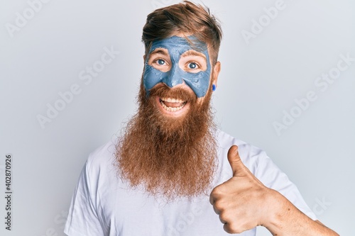 Young irish redhead man wearing facial mask smiling happy and positive, thumb up doing excellent and approval sign
