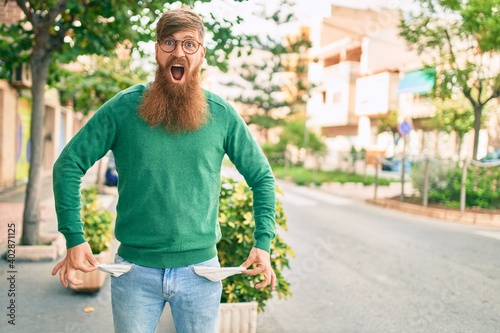 Young irish man with redhead beard surprised and showing his empty pockets jeans at the city.