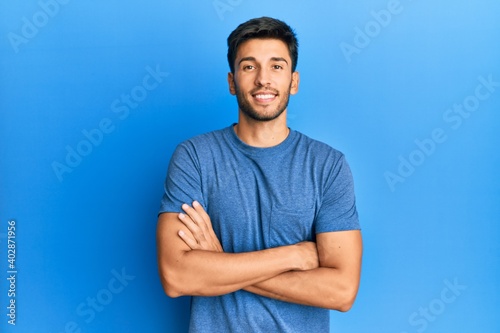 Young handsome man wearing casual tshirt over blue background happy face smiling with crossed arms looking at the camera. positive person.