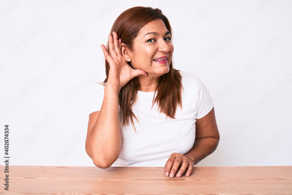 Middle age brunette hispanic woman wearing casual white tshirt sitting on the table smiling with hand over ear listening and hearing to rumor or gossip. deafness concept. Middle age brunette hispanic woman wearing casual white tshirt sitting on the table smiling with hand over ear listening and hearing to rumor or gossip. deafness concept.