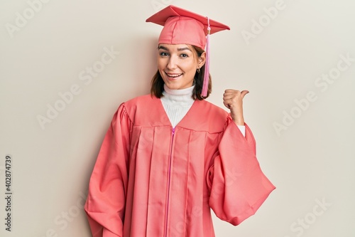 Young caucasian woman wearing graduation cap and ceremony robe smiling with happy face looking and pointing to the side with thumb up.