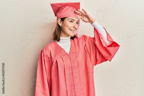 Young caucasian woman wearing graduation cap and ceremony robe very happy and smiling looking far away with hand over head. searching concept.