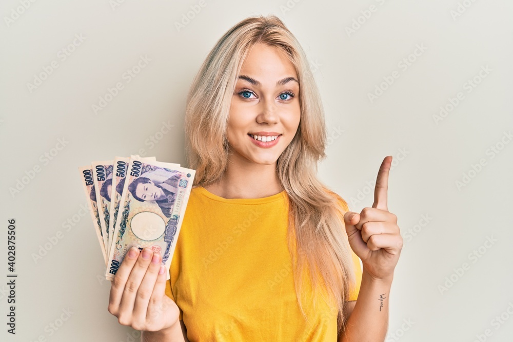 Beautiful caucasian blonde girl holding 5000 japanese yen banknotes smiling with an idea or question pointing finger with happy face, number one Beautiful caucasian blonde girl holding 5000 japanese yen banknotes smiling with an idea or question pointing finger with happy face, number one