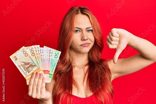 Young beautiful redhead woman holding norwegian krone banknotes with angry face, negative sign showing dislike with thumbs down, rejection concept