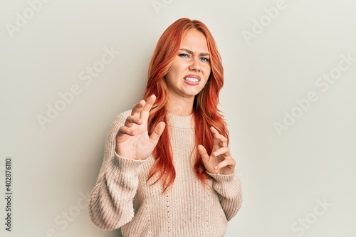 Young beautiful redhead woman wearing casual winter sweater disgusted expression, displeased and fearful doing disgust face because aversion reaction. with hands raised