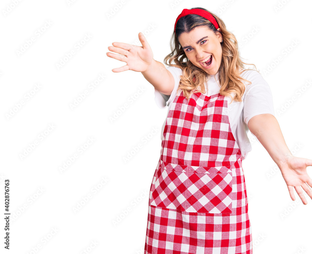 Young caucasian woman wearing apron looking at the camera smiling with open arms for hug. cheerful expression embracing happiness. Young caucasian woman wearing apron looking at the camera smiling with open arms for hug. cheerful expression embracing happiness.