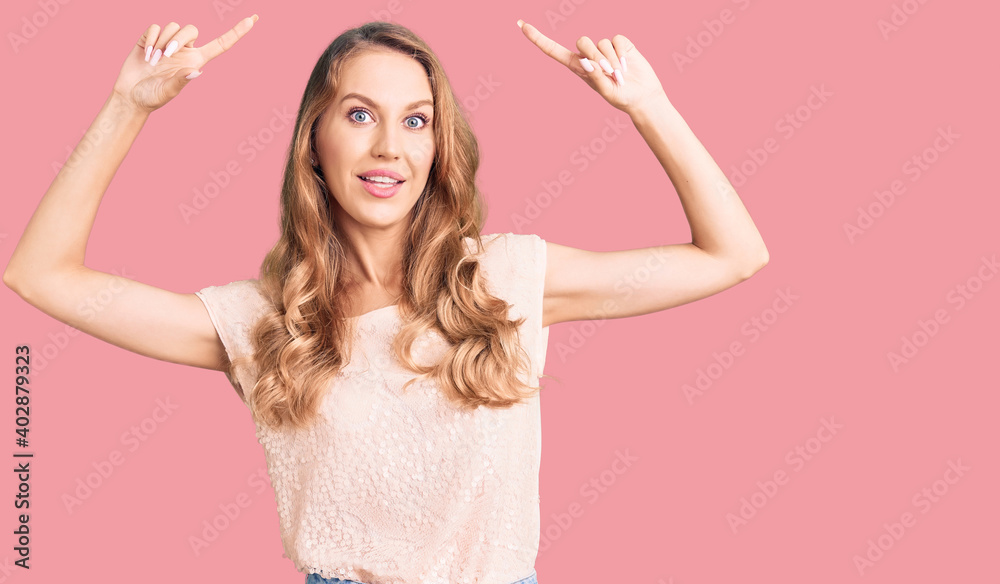 Young beautiful caucasian woman with blond hair wearing casual clothes smiling amazed and surprised and pointing up with fingers and raised arms. Young beautiful caucasian woman with blond hair wearing casual clothes smiling amazed and surprised and pointing up with fingers and raised arms.