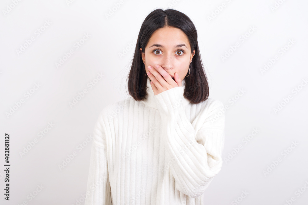 Emotional Young brunette woman wearing white knitted sweater against white background gasps from astonishment, covers opened mouth with palm, looks shocked at camera. Reaction concept Emotional Young brunette woman wearing white knitted sweater against white background gasps from astonishment, covers opened mouth with palm, looks shocked at camera. Reaction concept