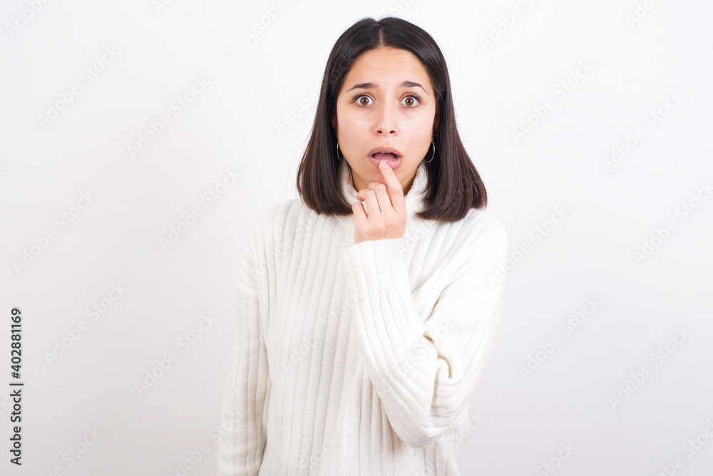 Nervous puzzled Young brunette woman wearing white knitted sweater against white background opens mouth from surprise, reacts on sudden news. Nervous puzzled Young brunette woman wearing white knitted sweater against white background opens mouth from surprise, reacts on sudden news.