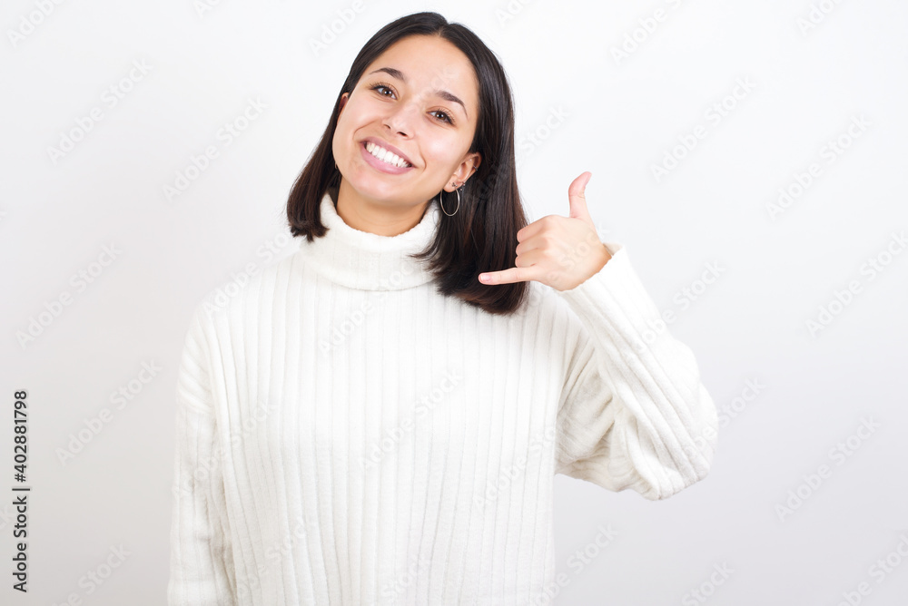 Young brunette woman wearing white knitted sweater against white background smiling doing phone gesture with hand and fingers like talking on the telephone. Communicating concepts. Young brunette woman wearing white knitted sweater against white background smiling doing phone gesture with hand and fingers like talking on the telephone. Communicating concepts.