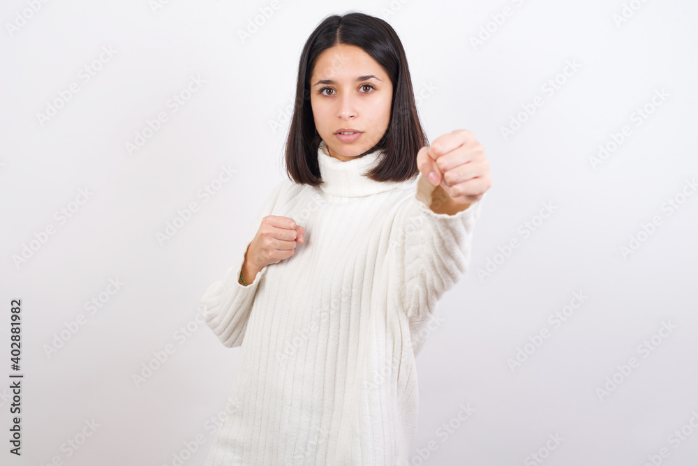 Young brunette woman wearing white knitted sweater against white background Punching fist to fight, aggressive and angry attack, threat and violence Young brunette woman wearing white knitted sweater against white background Punching fist to fight, aggressive and angry attack, threat and violence