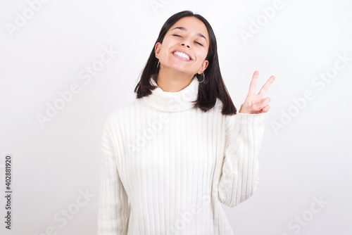 Young brunette woman wearing white knitted sweater against white background smiling with happy face winking at the camera doing victory sign. Number two.