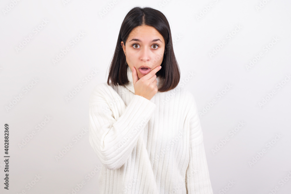 Young brunette woman wearing white knitted sweater against white background Looking fascinated with disbelief, surprise and amazed expression with hands on chin