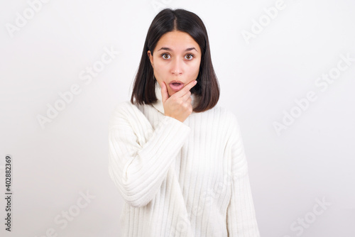 Young brunette woman wearing white knitted sweater against white background Looking fascinated with disbelief, surprise and amazed expression with hands on chin © Roquillo