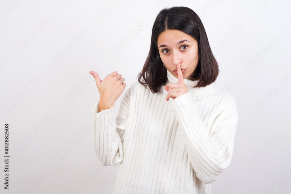 Young brunette woman wearing white knitted sweater against white background asking to be quiet with finger on lips pointing with hand to the side. Silence and secret concept. Young brunette woman wearing white knitted sweater against white background asking to be quiet with finger on lips pointing with hand to the side. Silence and secret concept.