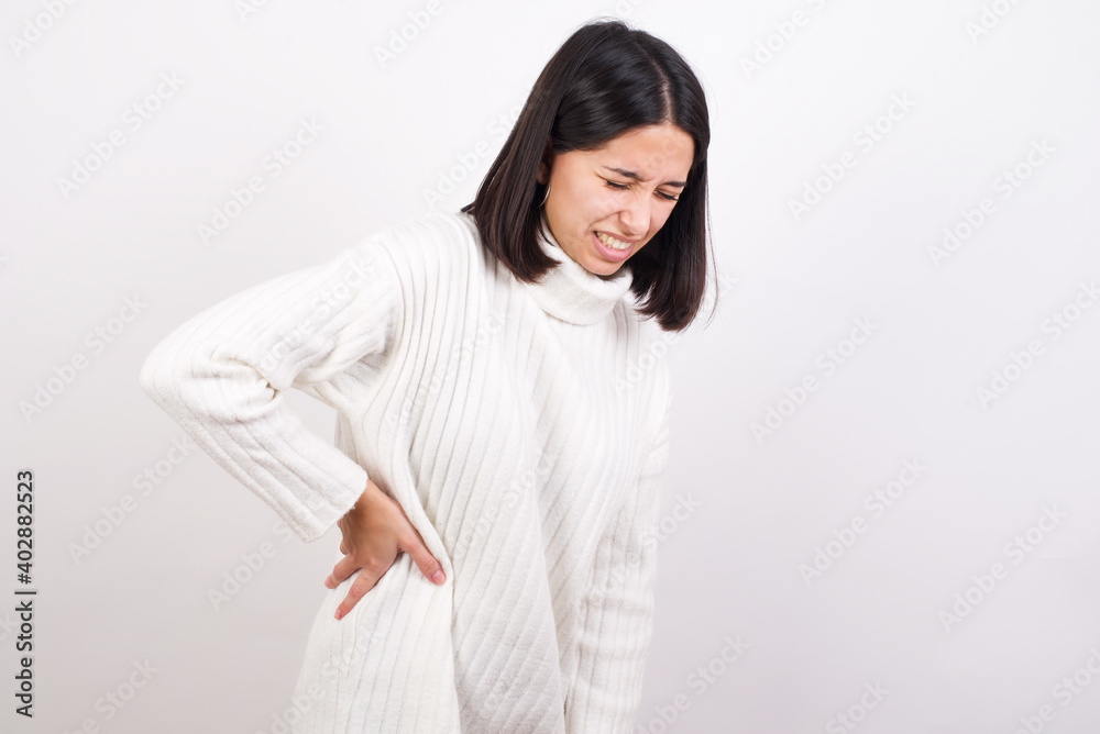 Young brunette woman wearing white knitted sweater against white background Suffering of backache, touching back with hand, muscular pain Young brunette woman wearing white knitted sweater against white background Suffering of backache, touching back with hand, muscular pain