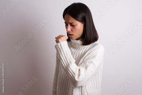 Young brunette woman wearing white knitted sweater against white background feeling unwell and coughing as symptom for cold or bronchitis. Healthcare concept.