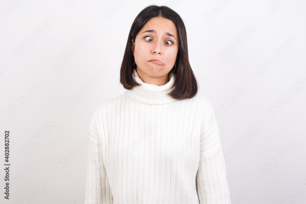 Fototapeta premium Young brunette woman wearing white knitted sweater against white background making grimace and crazy face, screaming out of control, funny lunatic expressing freedom and wild.