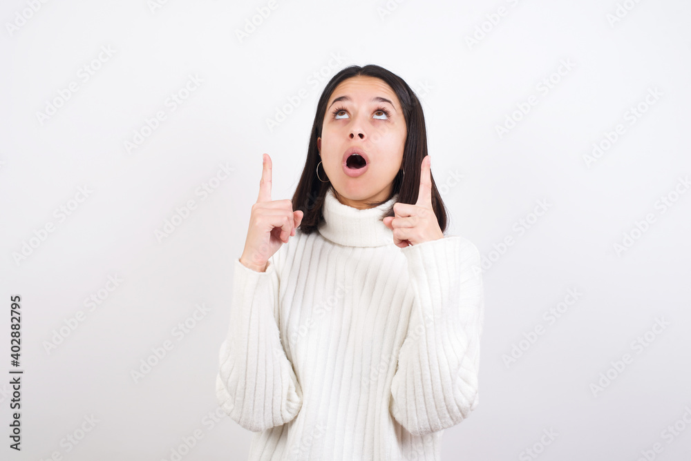 Young brunette woman wearing white knitted sweater against white background amazed and surprised looking up and pointing with fingers and raised arms. Young brunette woman wearing white knitted sweater against white background amazed and surprised looking up and pointing with fingers and raised arms.