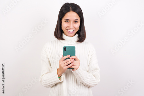 Young brunette woman wearing white knitted sweater against white background enjoys distant communication, uses mobile phone, surfs fast unlimited internet, has pleasant smile, makes shopping online,