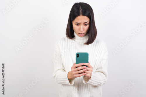 Focused Young brunette woman wearing white knitted sweater against white background use smartphone reading social media news, or important e-mail