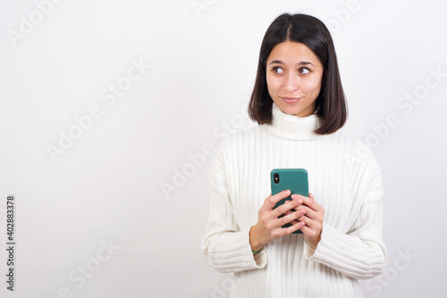 Young brunette woman wearing white knitted sweater against white background holds telephone hands reads good youth news look empty space advert