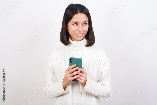 Young brunette woman wearing white knitted sweater against white background hold telephone hands read good youth news look empty space advert